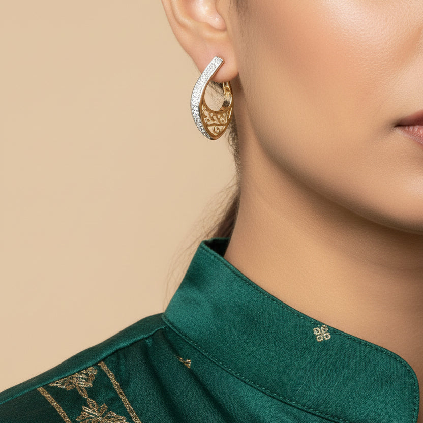 A pair of gold crescent-shaped hoop earrings with silver pavé trim and an openwork filigree center, displayed on a white card against a textured gray background.