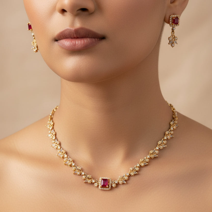 Gold-toned jewelry set displayed on a black velvet mannequin bust, featuring a necklace with a central red gemstone and matching earrings, photographed outdoors on a tiled ledge with a garden and gray wall in the background.